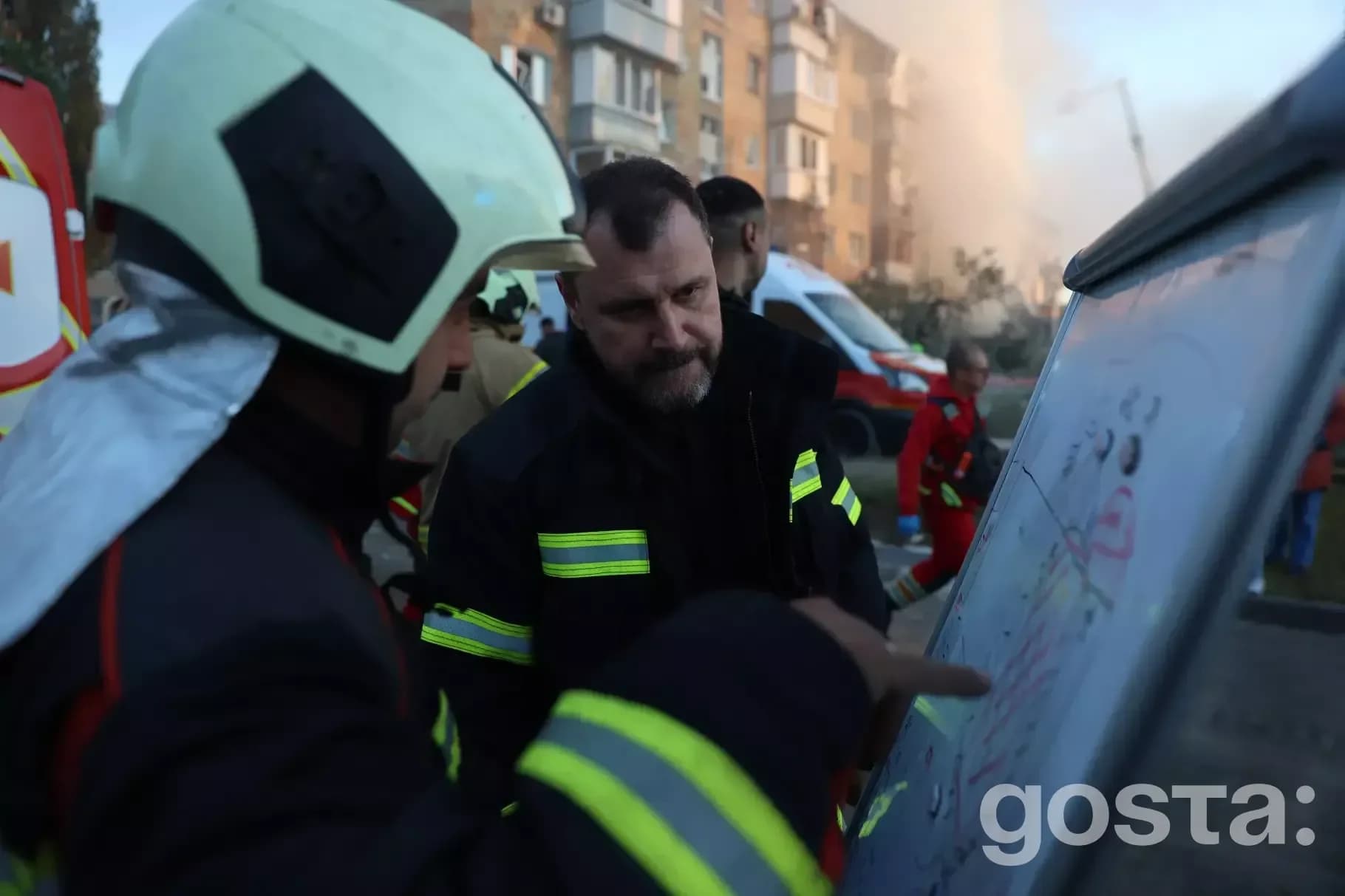 На зображенні рятувальники обговорюють план дій на тлі пошкоджених будівель після обстрілу в Україні.