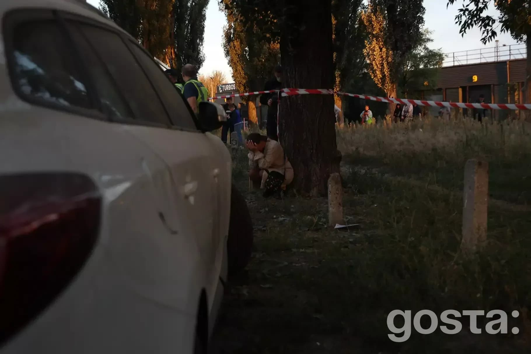На зображенні бачимо місце події, оточене стрічкою, з поліцейськими і людьми біля дерева у вечірній час.