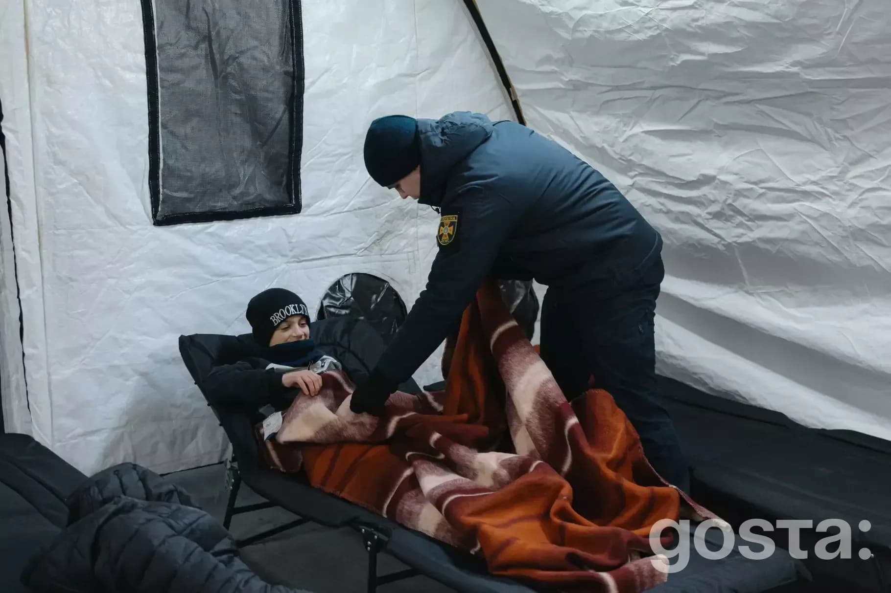 Рятувальник ДСНС України вкриває дитину ковдрою в наметовому містечку Троєщини після обстрілу.
