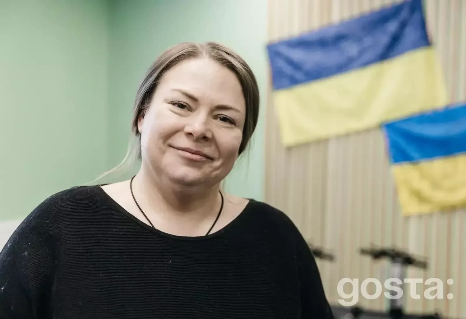 На фото жінка в приміщенні з двома українськими прапорами на стіні.