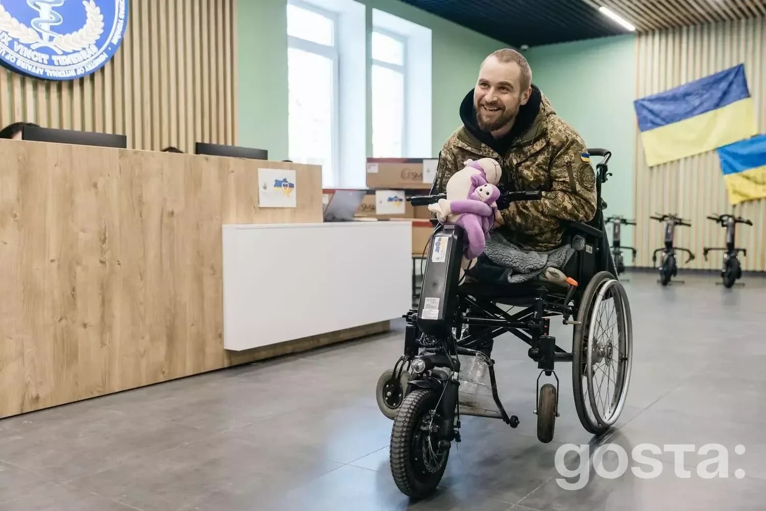 Чоловік у військовій формі на інвалідному візку з електроприводом, у приміщенні медичного закладу, посміхається.