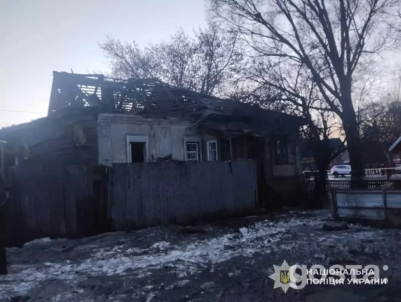 Напівзруйнований будинок після обстрілу в Україні; пошкоджений дах, вікна, навколо зимові дерева, логотип поліції.