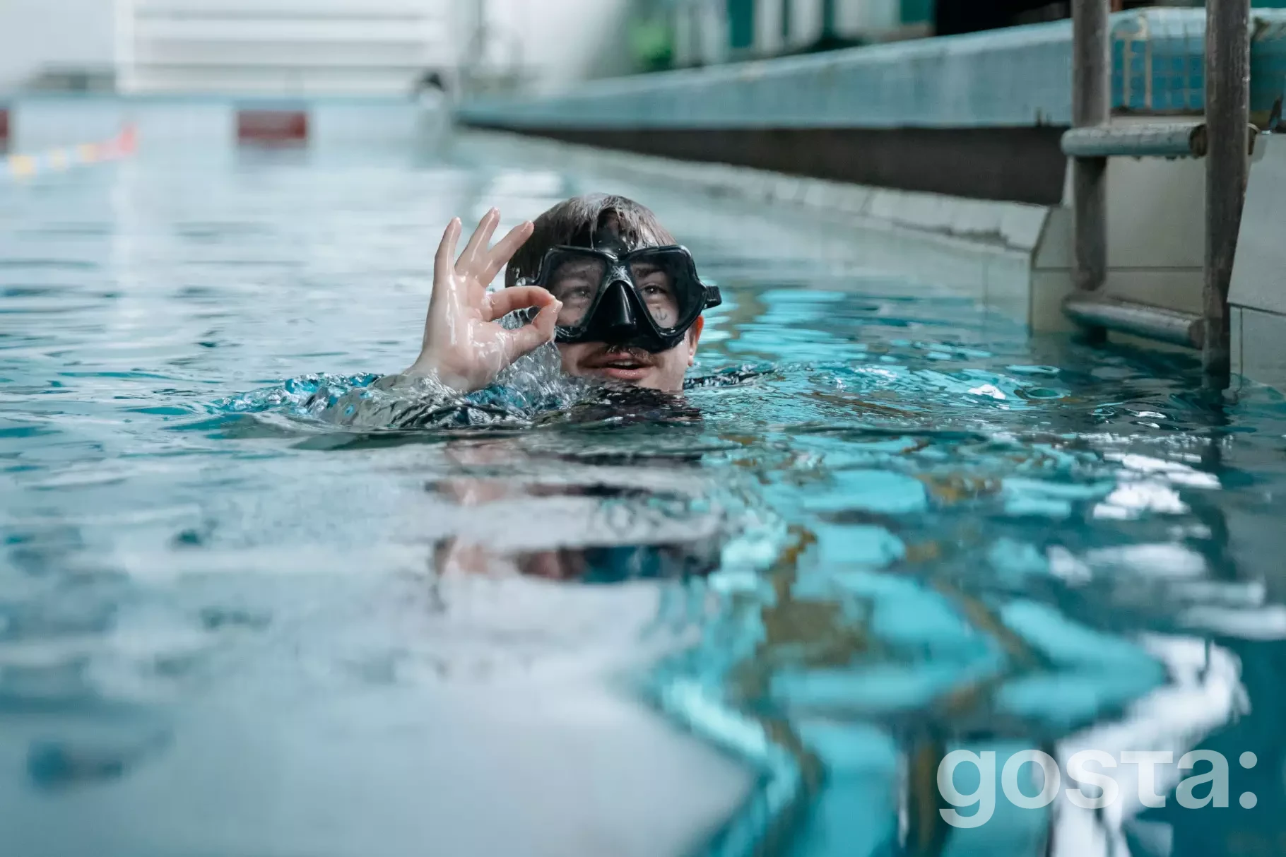На зображенні людина в басейні з обладнанням для дайвінгу демонструє жест "OK" під час реабілітації.