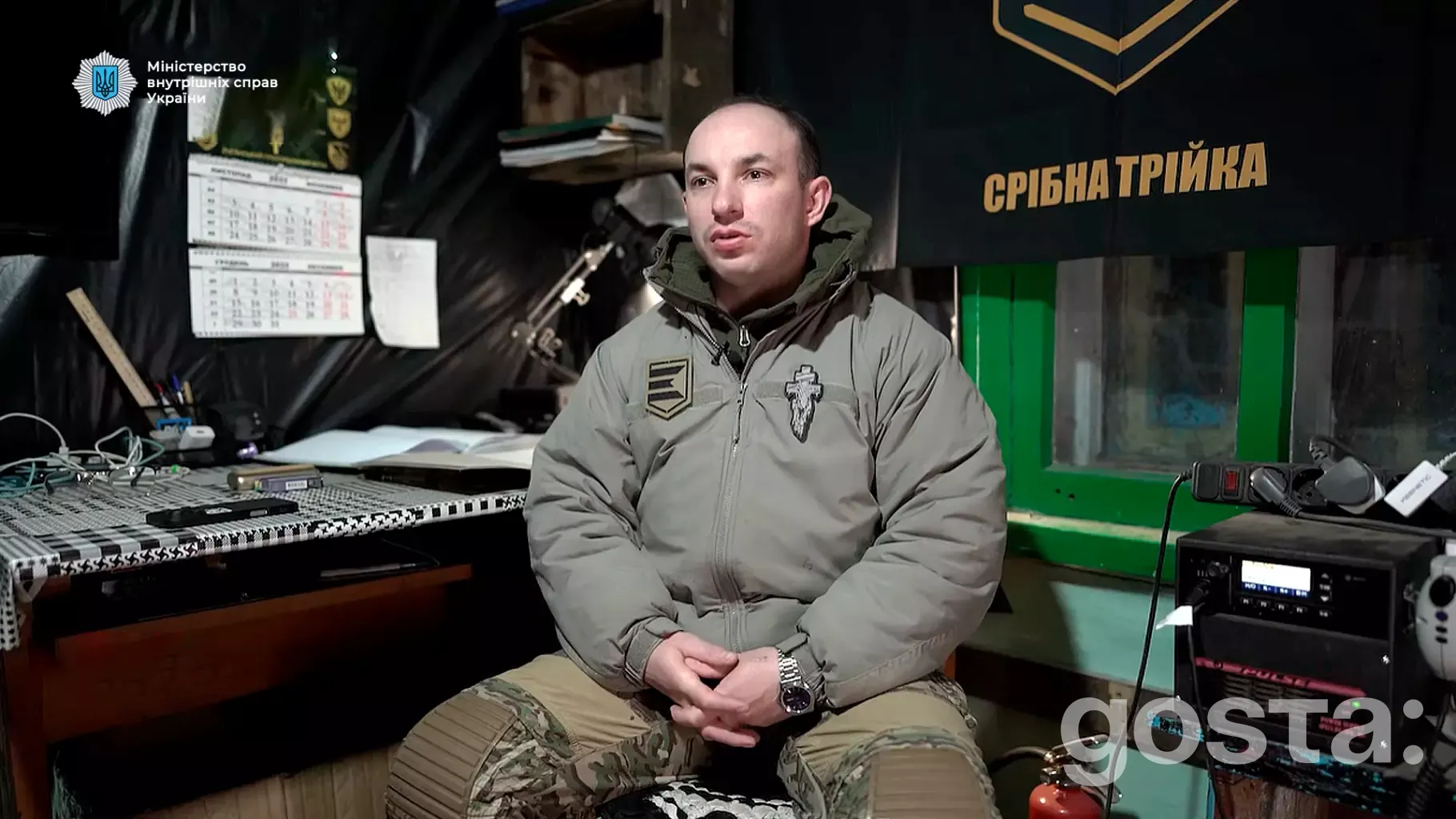 На зображенні чоловік у військовій формі сидить у кімнаті з військовою символікою, розповідає про евакуацію.