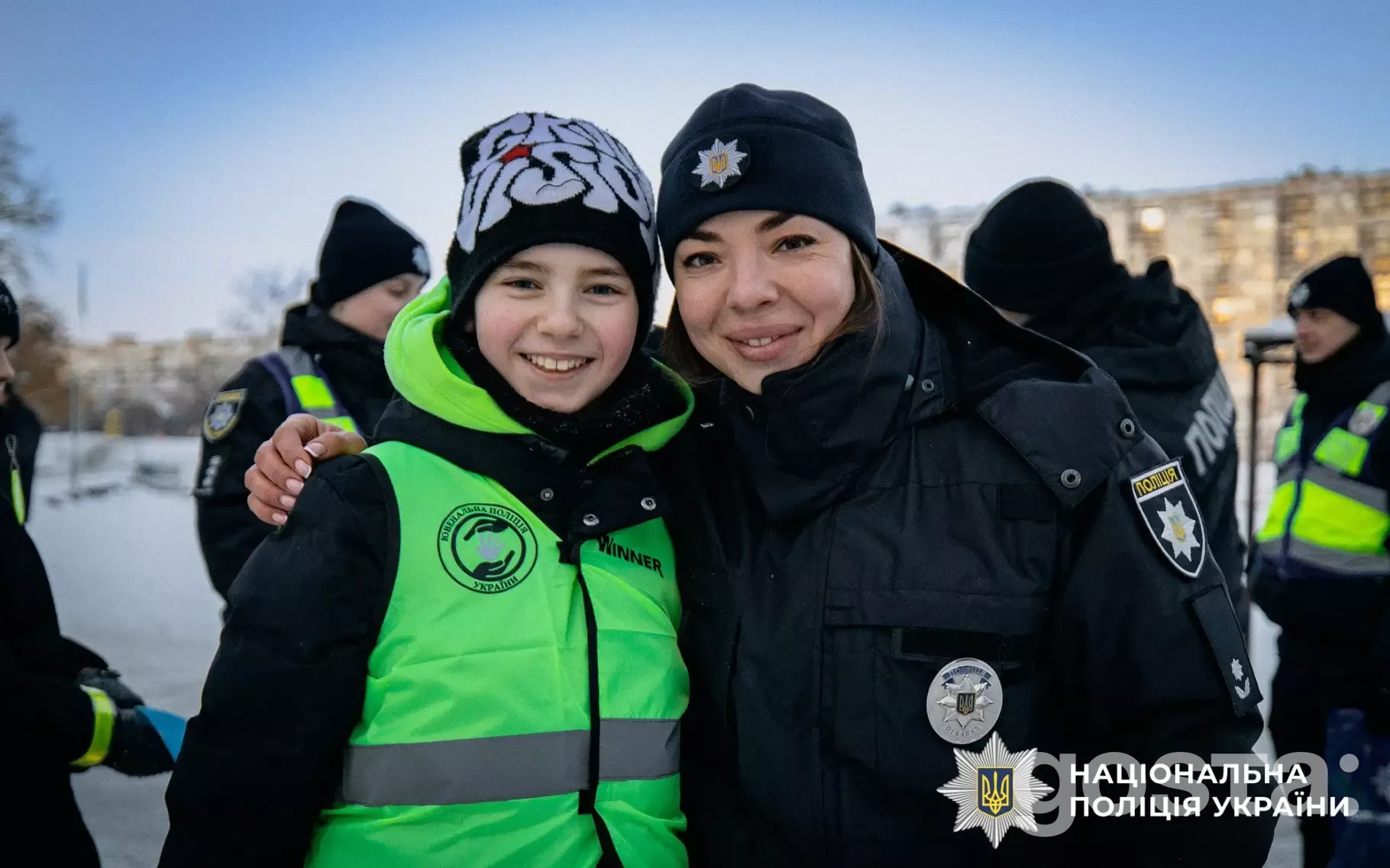 На зображенні усміхнені ювенальні поліцейські з дитиною в зимовий час, Одягнені у форму зі світловідбивальними елементами.