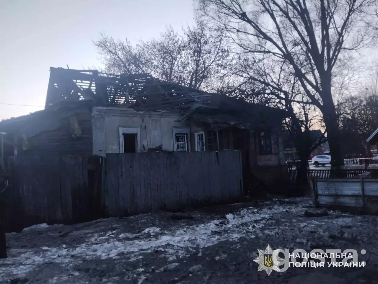 Напівзруйнований будинок після обстрілу в Україні; пошкоджений дах, вікна, навколо зимові дерева, логотип поліції.