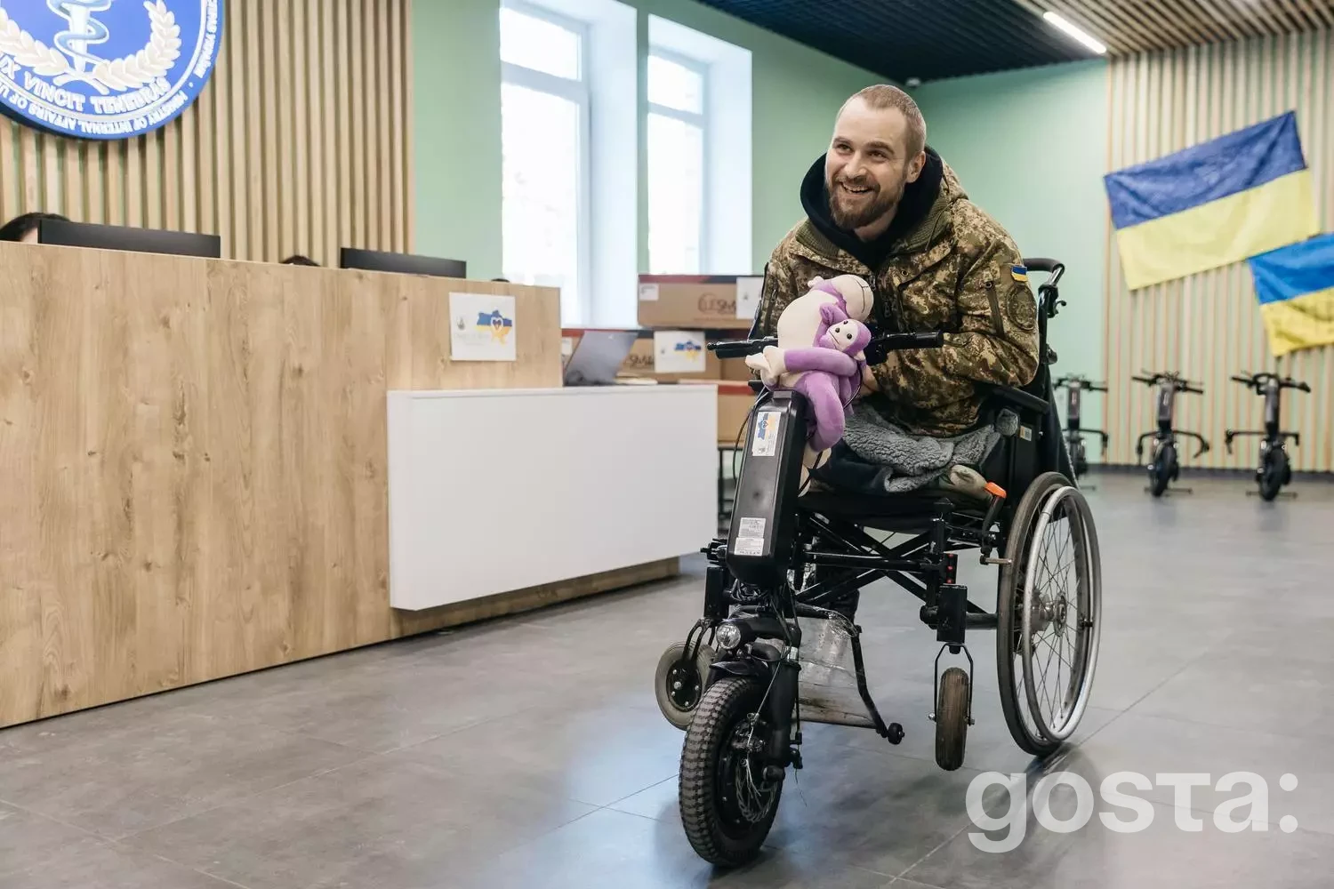 Чоловік у військовій формі на інвалідному візку з електроприводом, у приміщенні медичного закладу, посміхається.