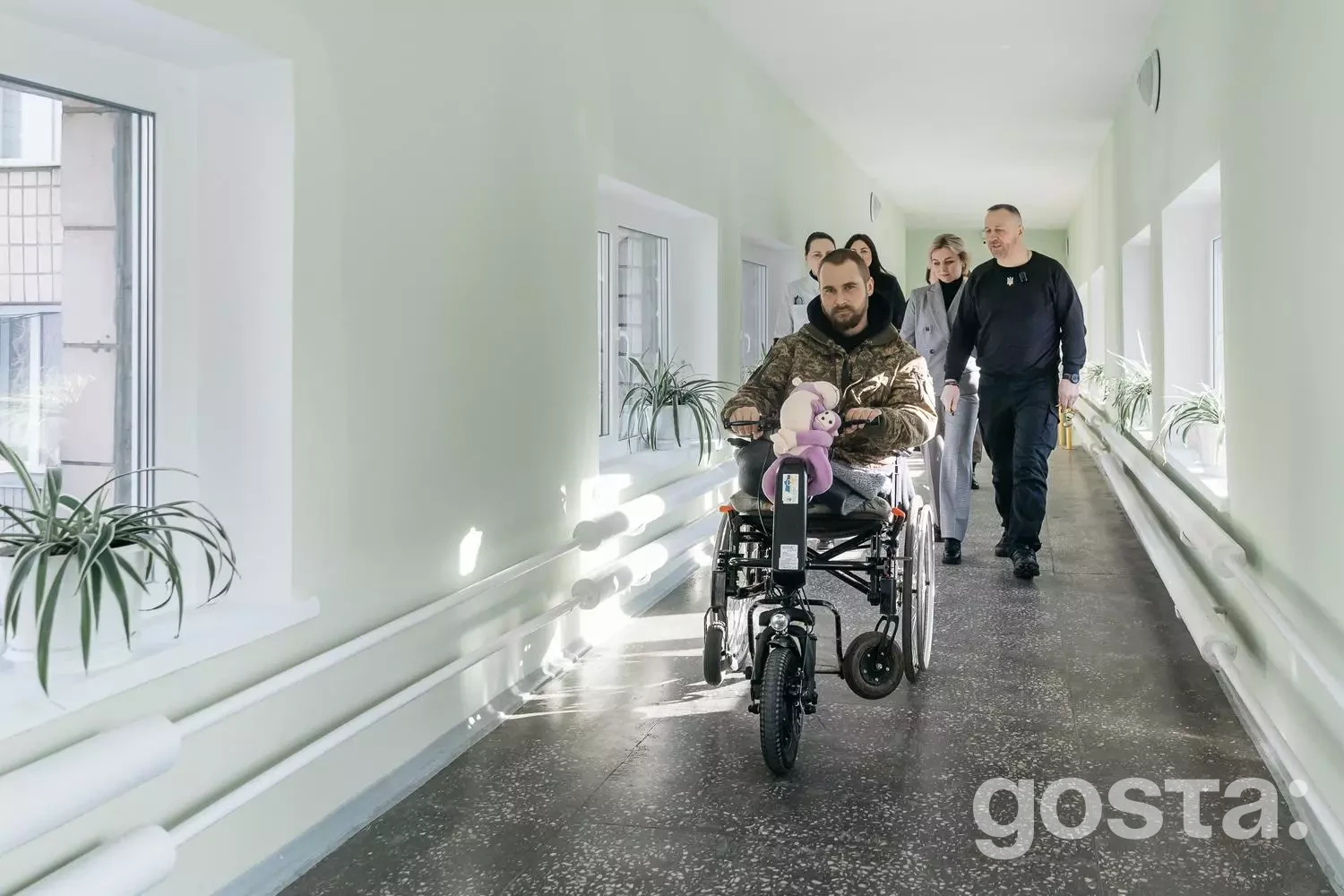 Чоловік на електричному інвалідному візку в коридорі госпіталю, супроводжуваний працівниками.