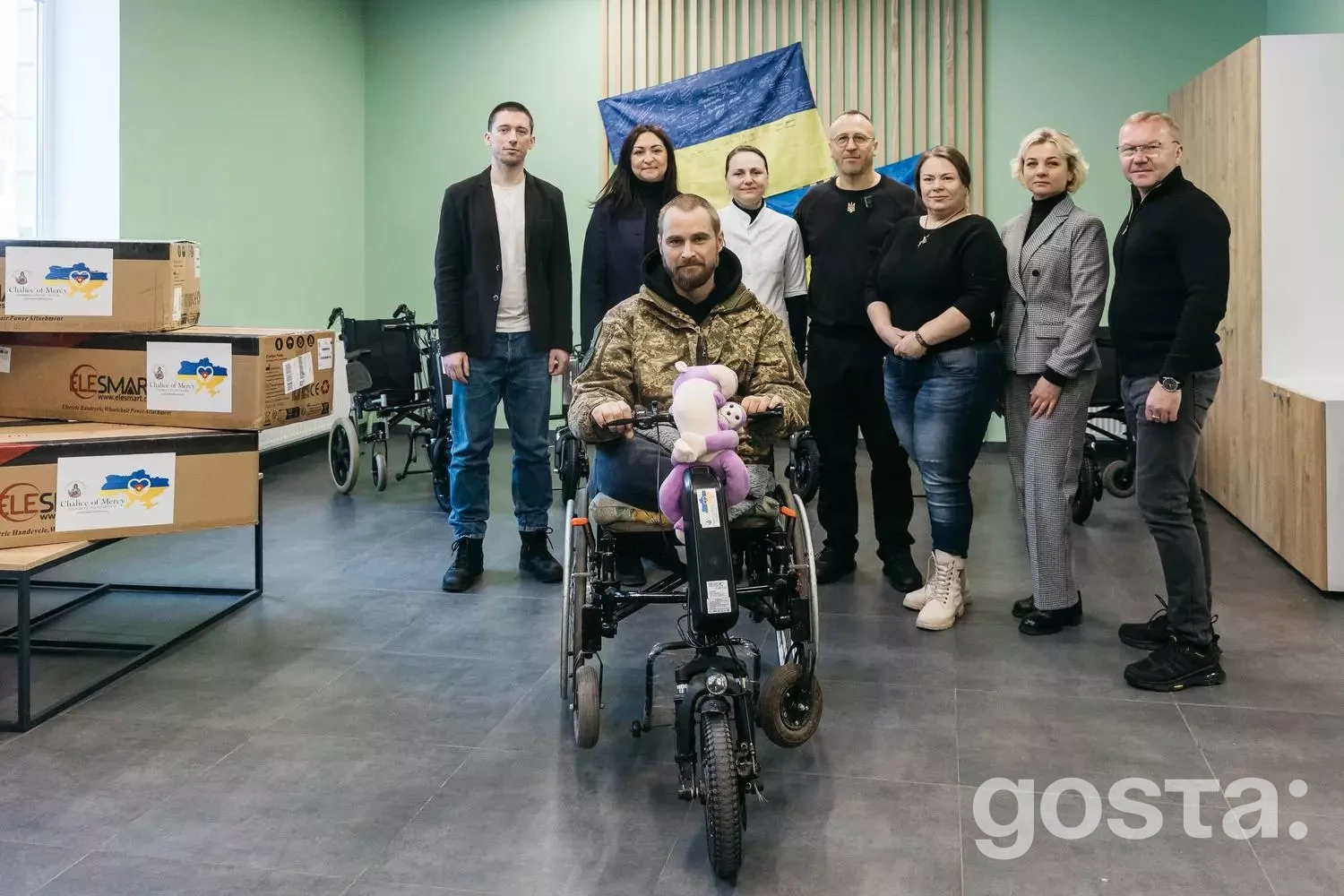 Група людей у приміщенні; поранений ветеран у кріслі з електроприводом, прапор України, благодійна підтримка.