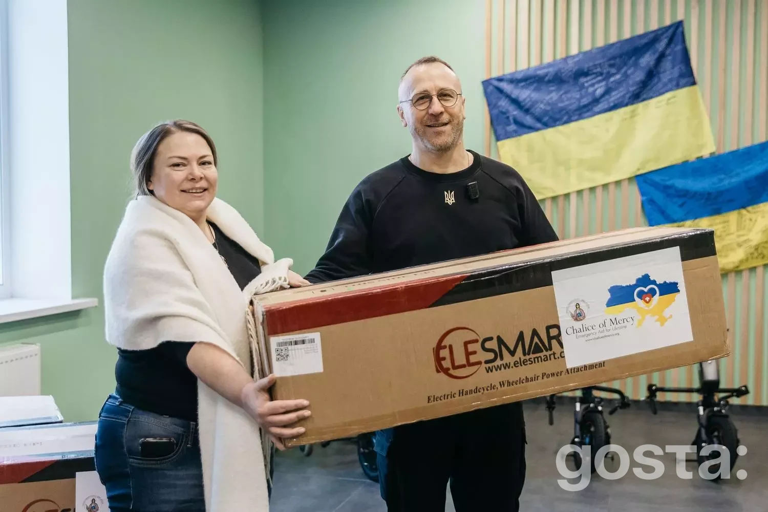 Я не можу впізнати людей на фото, але можу описати його.  Двоє людей тримають коробку з електроприводом для крісла у цент...