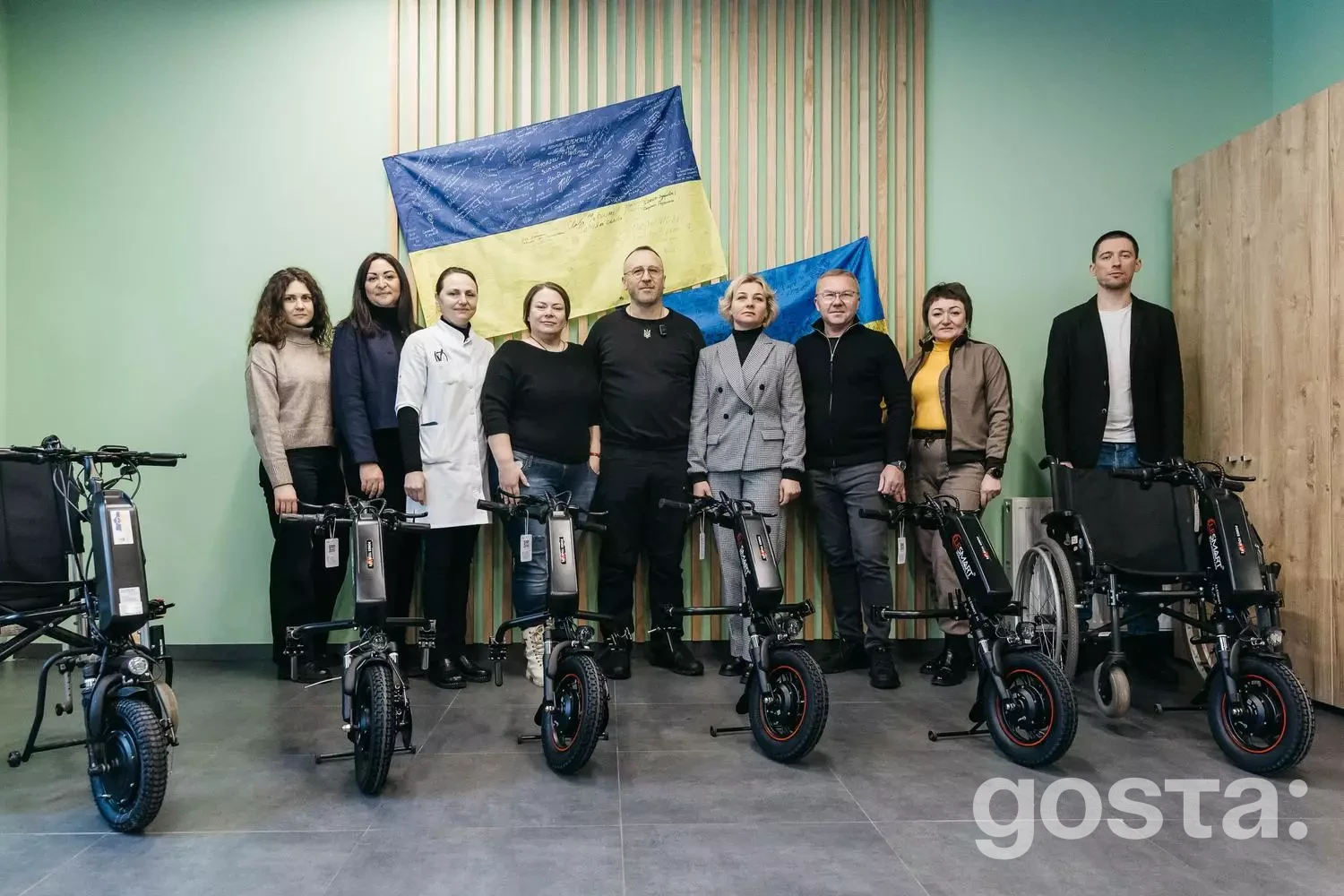 Група людей з електричними приводами для інвалідних візків, прапор України на фоні, захід у медичному центрі МВС.