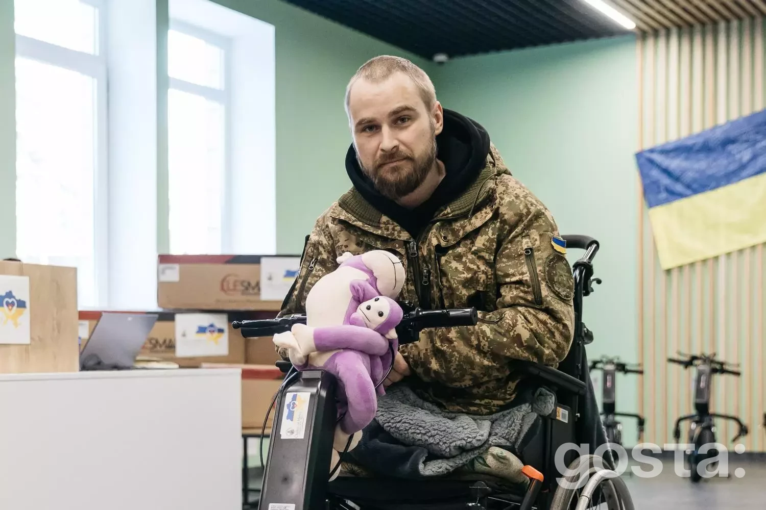 На зображенні: ветеран у камуфляжі сидить у візку з електроприводом, тримаючи м'яку іграшку, у приміщенні медичного закладу.