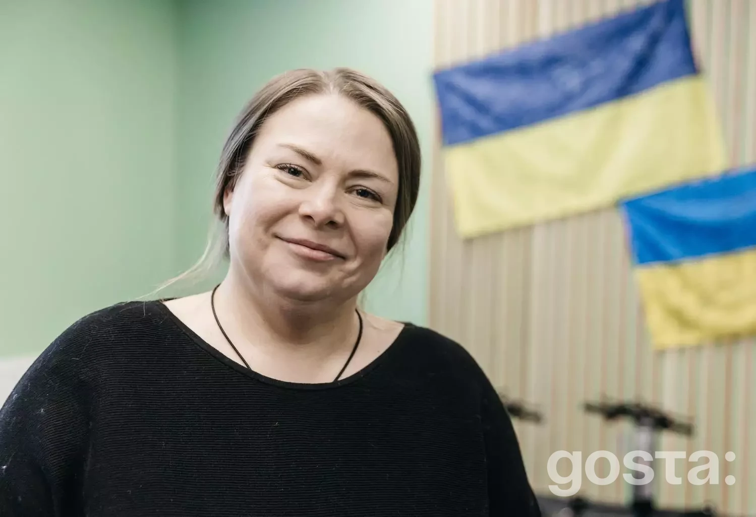 На фото жінка в приміщенні з двома українськими прапорами на стіні.