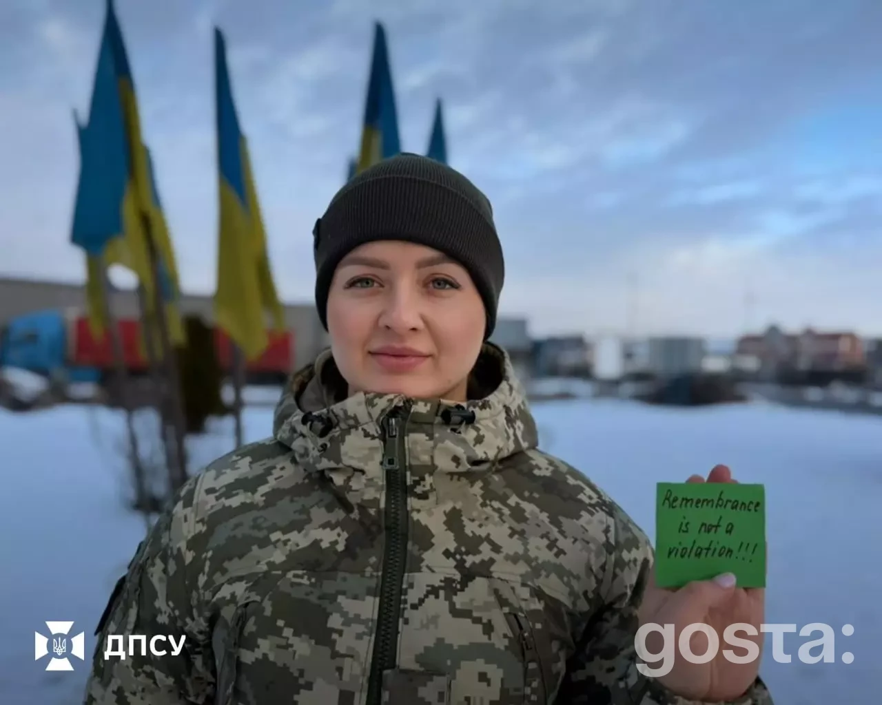 На фото військовослужбовець тримає табличку "Remembrance is not a violation" на фоні прапорів України.