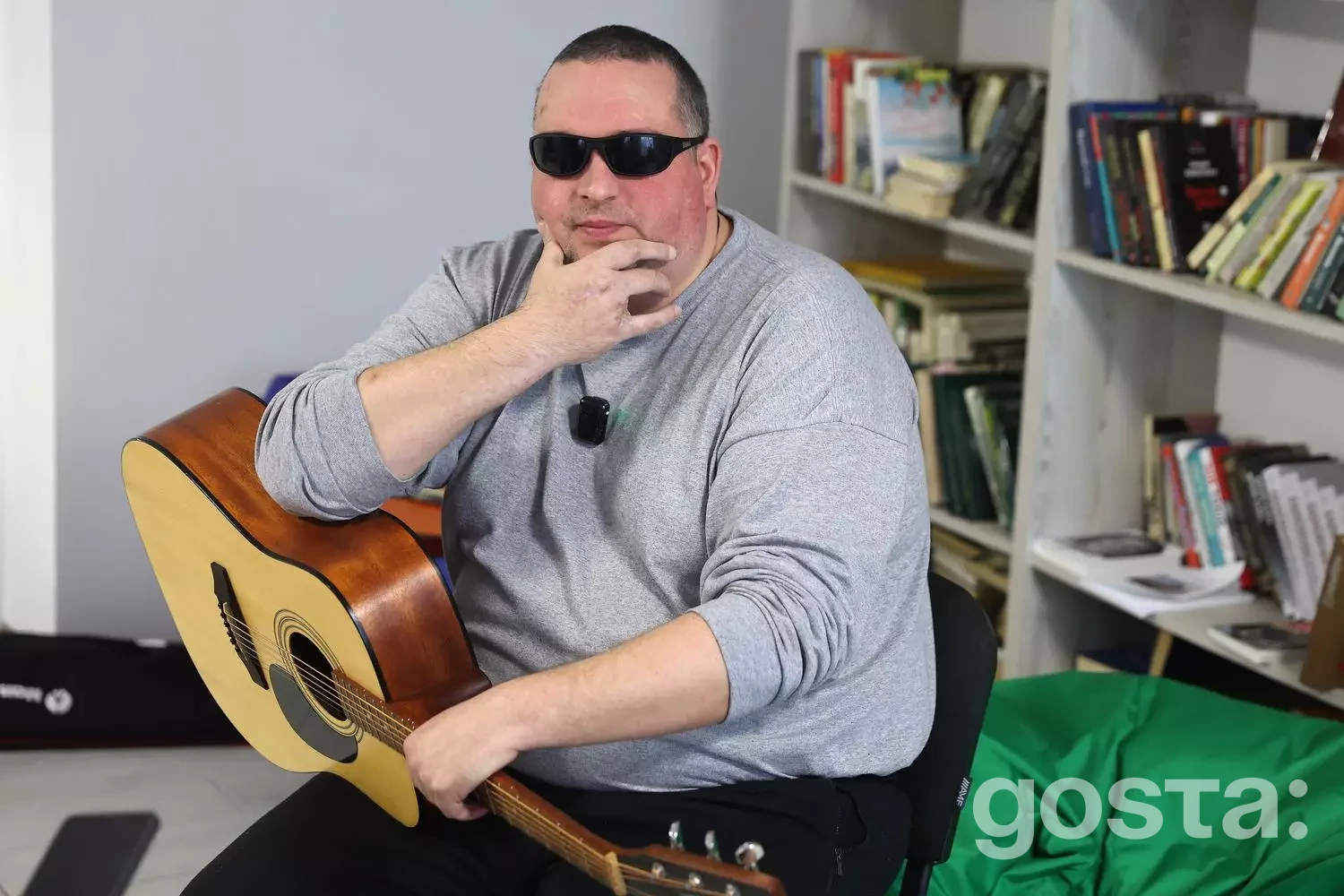 На зображенні чоловік у темних окулярах тримає гітару в реабілітаційному центрі, поруч полиці з книгами.