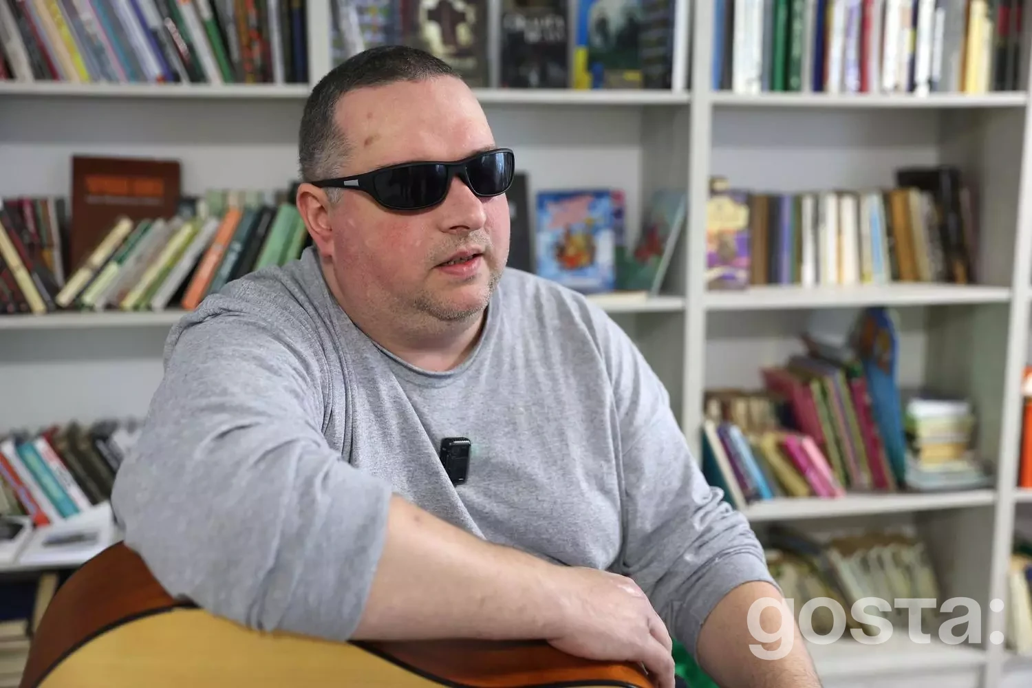 На зображенні чоловік у сонцезахисних окулярах тримає гітару в бібліотеці, долаючи виклики реабілітації.