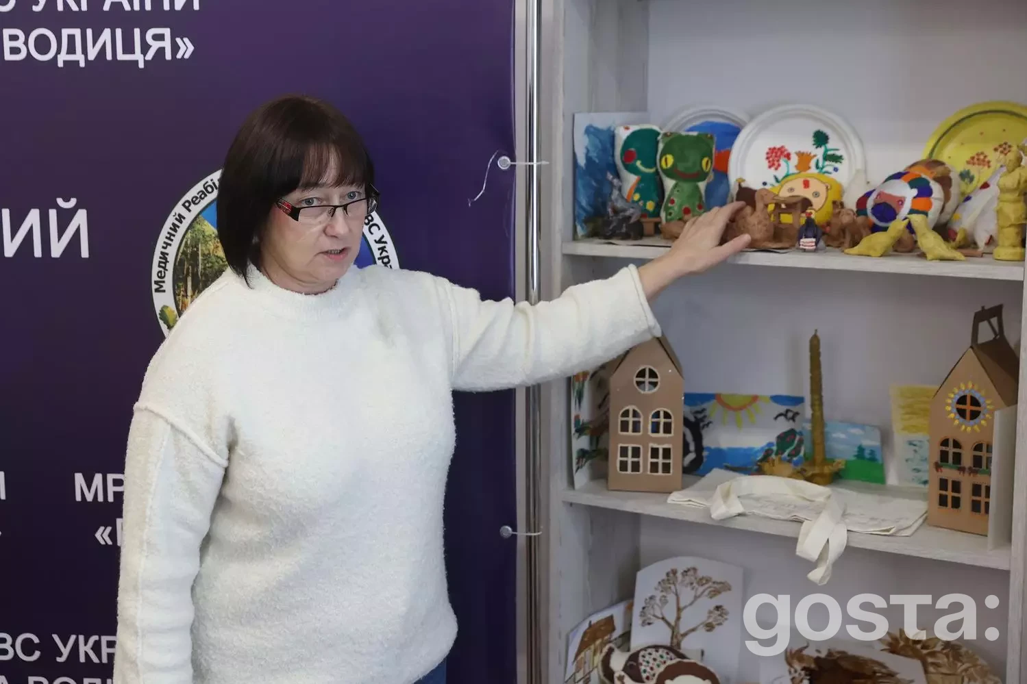 Волонтерка у білому светрі показує ремісничі вироби в медичному реабілітаційному центрі.