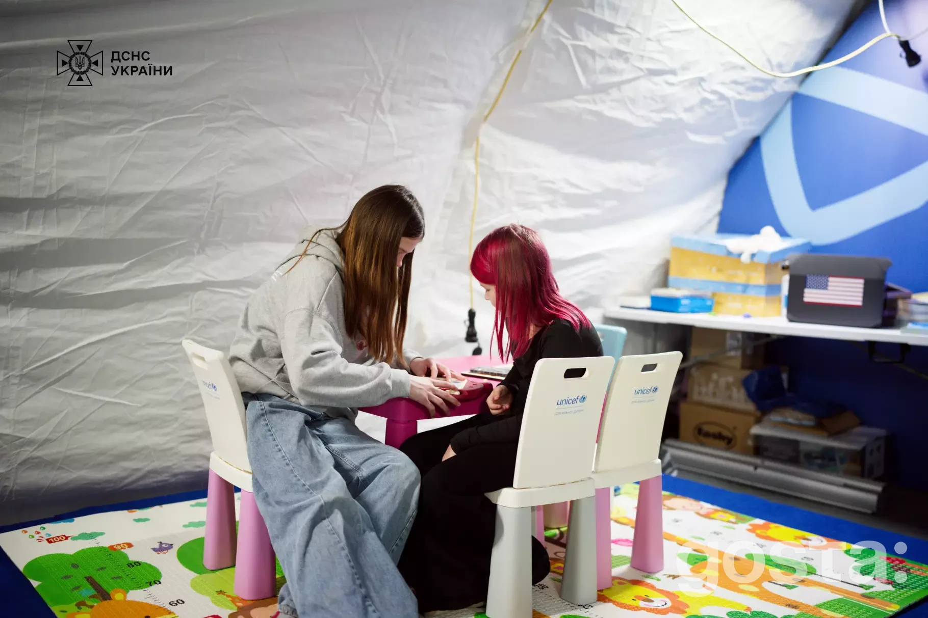 На фото двоє дітей, що сидять за столом у мобільному пункті ДСНС та ЮНІСЕФ для психосоціальної підтримки.